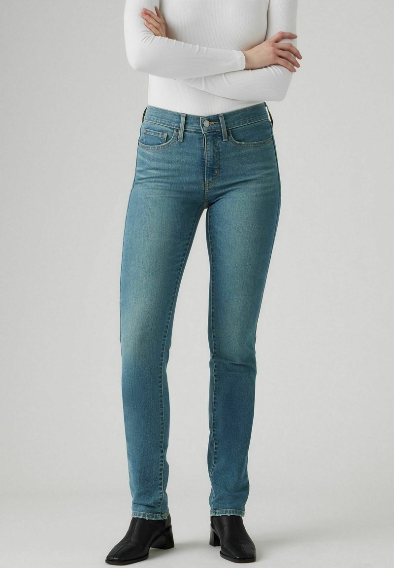 Personne portant un jean slim bleu clair, des bottines noires et un haut blanc à manches longues, bras croisés.
