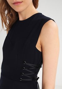 Marindress i marinblått med en figursydd design, slät textur och sidoknytningar med svarta band trätt genom metallöglor.