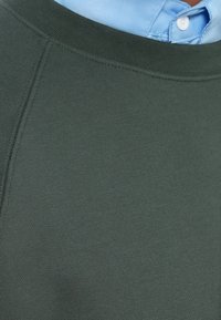 Detailní záběr na tmavě zelený svetr nošený přes světle modrou košilovou košili, přičemž u límce je vidět bílý knoflík.
