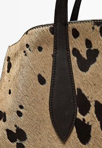 Borsa a tracolla in pelle di mucca tan con macchie scure, dotata di manici e cuciture in pelle nera. La texture è morbida con una finitura elegante.
