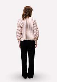 Vrouw met haar rug naar de kijker gekeerd, in een cremekleurige blouse met lange mouwen, gekleed in roze en paarse verticale strepen en zwarte broek met wijde pijpen, tegen een witte achtergrond.