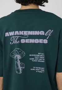 T-shirt vert foncé avec un texte graphique indiquant "Éveil des Sens" et une illustration de champignon en blanc. Coupe décontractée et col rond.