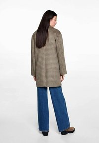 Rutet frakke i beige og brun stof, lange ærmer, afslappet pasform, parret med brede blå jeans og brune ankelsko.
