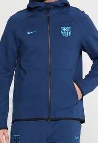 Sweat à capuche zippé bleu marine avec une coupe ajustée, arborant un grand logo FC Barcelone et une marque Nike en bleu clair. Comprend des poches avant et une capuche.