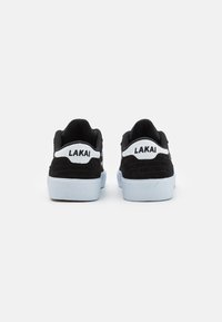 Black suede sneakers with a white logo, featuring a low-top design and light blue rubber sole, showcasing a textured back panel.