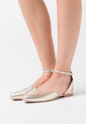 Pieds portant des chaussures plates dorées métalliques avec des brides de cheville et de petites boucles sur un fond blanc.