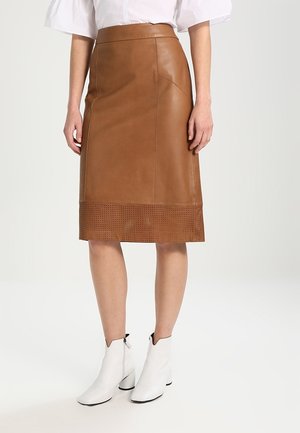 Femme portant une jupe en cuir marron longueur genoux avec ourlet perforé, une blouse blanche et des bottines blanches, debout devant un fond uni.