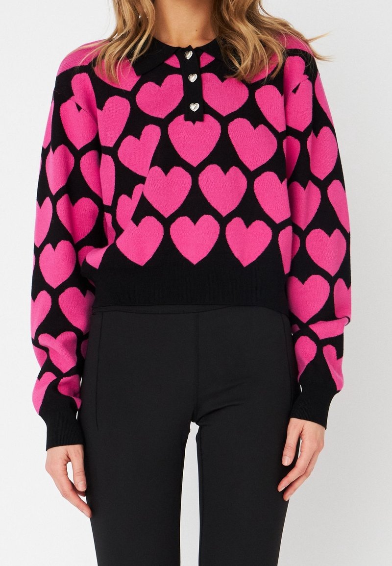 Femme portant un pull noir à manches longues avec des motifs de cœurs rose vif et un col noir orné de trois boutons en forme de cœur.