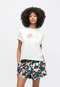 Svetlokrémové tričko s vyhrnutými rukávmi a gradientným logom "new balance"; v kombinácii s farebnými krátkymi nohavicami s motívom v modrej, žltej a červenej farbe.