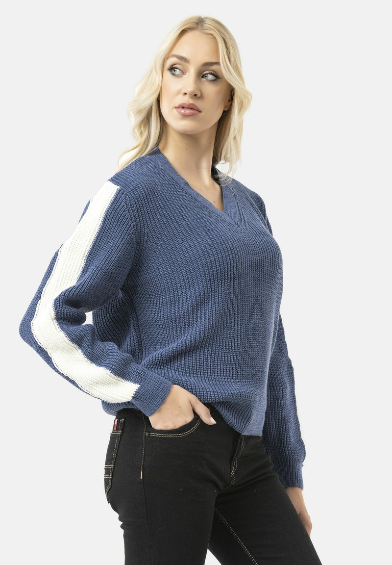 Blauwe gebreide trui met een V-hals, lange mouwen met witte streepaccenten en een gestructureerde afwerking. Gepaard met zwarte jeans.