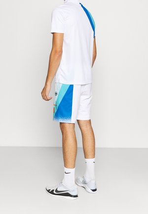 Mand iført hvidt sportstøj med blå geometriske mønstre på shorts, stående mod en ensartet lys baggrund, med ryggen mod kameraet.
