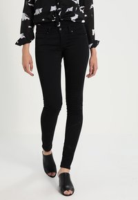 Jeans negros ajustados con un acabado suave, que cuentan con un diseño clásico de cinco bolsillos y una cintura de altura media, combinados con zapatos de tacón abiertos negros.