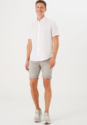 Camisa blanca de manga corta y botones, hecha de un tejido ligero, combinada con pantalones cortos gris claro arremangados y zapatos deportivos grises.