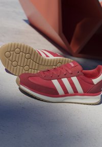 Baskets rouges Adidas avec des bandes blanches, des semelles et des lacets beige, posées sur un sol gris clair avec un objet géométrique marron en arrière-plan.