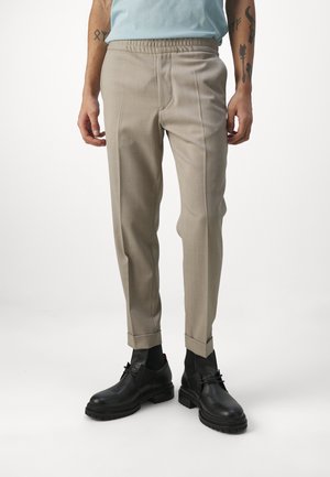 Partie inférieure d'une personne portant un pantalon beige slim-fit avec des ourlets retroussés et des bottines en cuir noir à lacets, debout sur un fond blanc.