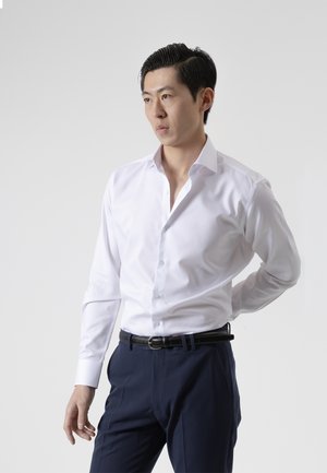 Joven con el pelo peinado hacia atrás, vistiendo una camisa blanca y pantalones azul marino, de pie con una mano detrás de la espalda, mirando de lado.