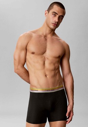 Gespierde man met kort haar die zwarte Calvin Klein boxershorts draagt met een verloop aan de tailleband, staand tegen een effen lichtgrijze achtergrond.