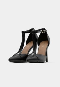 Μαύρες γόβες με λουράκι Τ, μυτερά δάχτυλα και αγκράφες στον αστράγαλο, τοποθετημένες η μία δίπλα στην άλλη σε λευκό φόντο.