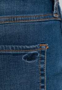 Blue denim jeans featuring a back pocket with an embroidered logo, orange stitching accentuating the seams, and a smooth texture.