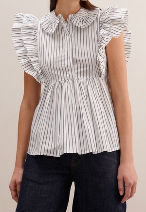 Hvid ærmeløs bluse med lodrette sorte striber, flæsekant ved krave og skuldre, parret med mørkeblå jeans.