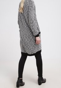 Dlouhý cardigan v černobílém textu s volným střihem, zdobený černým lemem. V kombinaci s černými legínami a zdobenými kotníkovými botami.