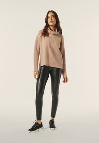 Lichtroze gebreid trui met een col, gecombineerd met glanzende zwarte leggings. Het model draagt zwarte sneakers met witte zolen.