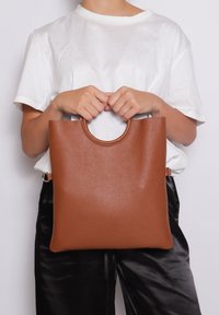 Borsa tote in pelle marrone con superficie testurizzata, forma rettangolare e manico circolare sulla parte superiore, tenuta da una persona in camicia bianca.
