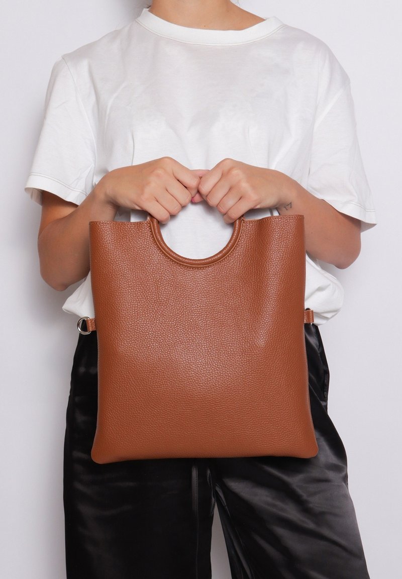 Borsa tote in pelle marrone con superficie testurizzata, forma rettangolare e manico circolare sulla parte superiore, tenuta da una persona in camicia bianca.