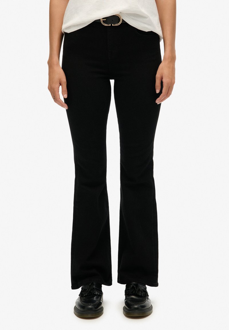 Jean negros de campana con una textura suave, ajuste de cintura alta y cinturón con hebilla dorada. Combinados con zapatos negros. Sin patrones o acentos visibles.