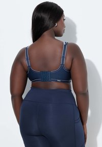 Marineblauwe sportbeha met verstelbare bandjes, haak- en oogsluiting en contrasterende stikseldetails; gecombineerd met marineblauwe leggings met hoge taille.