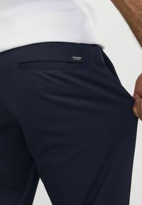 Uomo che indossa pantaloni blu navy Jack & Jones, tirando il tessuto vicino alla tasca posteriore mentre indossa una camicia bianca.