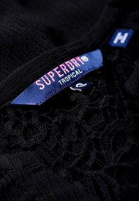 Tela negra con textura de encaje, con una etiqueta azul que dice "SUPERDRY TROPICAL" en rosa y blanco. Diseño cosido con patrones sutiles.