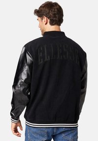 Black jacket with leather sleeves, "ELLESSE" embroidered on the back. Ribbed collar and cuffs, white stripe detailing at the hem.