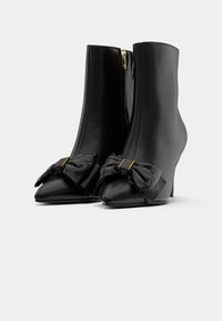 Zwarte leren enkelboots met spitse neuzen. Heeft een ritssluiting en een grote strikdetail aan de voorkant met een gouden accent.