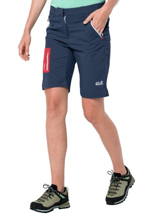 Personne portant un short de randonnée bleu marine avec des poches et des chaussures de randonnée, debout avec une main dans la poche et une jambe légèrement avancée.