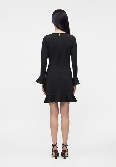 Femme aux longs cheveux foncés portant une robe noire à manches longues avec des volants aux poignets et à l'ourlet, ainsi que des sandales noires à talons, debout de dos.