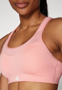 Reggiseno sportivo rosa chiaro con una superficie liscia, spalline larghe e scollatura rotonda, con un logo bianco sulla fascia.