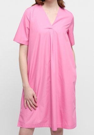 Donna che indossa un vestito rosa largo al ginocchio con maniche corte e scollo a V, in piedi su uno sfondo chiaro e uniforme.