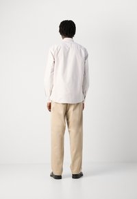 Camicia a maniche lunghe di colore chiaro a righe con polsini abbottonati, abbinata a pantaloni beige larghi e scarpe nere, vista da dietro.