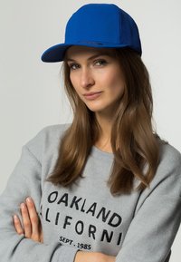 Királykék baseball sapka szerkezeti elülső résszel, hálós oldalakkal és ívelt karimával, egy szürke pulóverrel viselve, fekete felirattal.