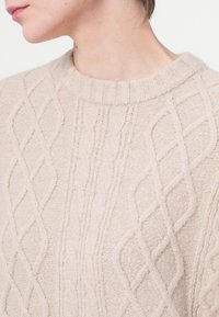 Pulover tricotat de culoare bej deschis, cu un design texturat de tip cablu, având guler rotund ribetat și un material moale, pufoș.