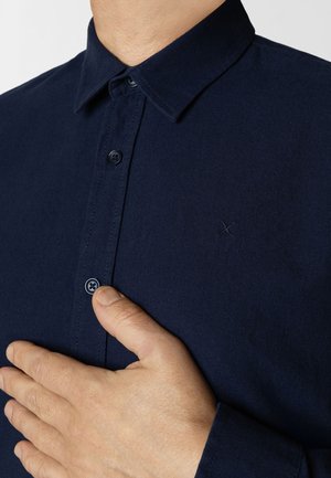 Marineblå skjorte med knapper lavet af stof med en glat tekstur; har en standardkrave og et lille broderet logo på brystet.