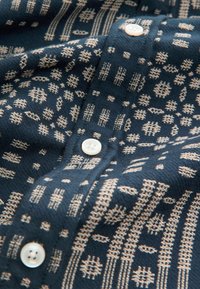 Tumesinine kangas särgiga, millel on kreemjad geomeetrilised mustrid; sisaldab nuppe ja tekstuurset kootud pinnast. Mustrites on jooned ja punktid üle pinna.