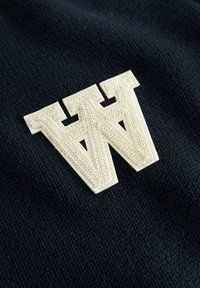 Tissu en maille bleu marine avec un grand logo "W" blanc texturé et bien en évidence. Détails fins des coutures et du motif tissé visibles.