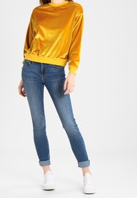 Sudadera de terciopelo amarillo con mangas largas y puños acanalados, combinada con jeans skinny de mezclilla azul y zapatillas blancas.