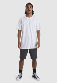 Camiseta blanca de algodón de manga corta, con corte relajado, combinada con pantalones cortos gris oscuro. El modelo lleva zapatillas negras y calcetines negros.