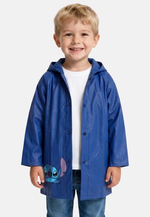 Jeune garçon souriant en imperméable bleu avec un motif de dessin animé Stitch, chemise blanche et jean, debout devant un fond uni clair.