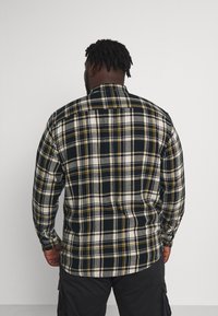 Svart, vit och gul rutmönstrad flanellskjorta med lång ärm och knapplister vid ärmsluten. Har krage och rundad fåll.