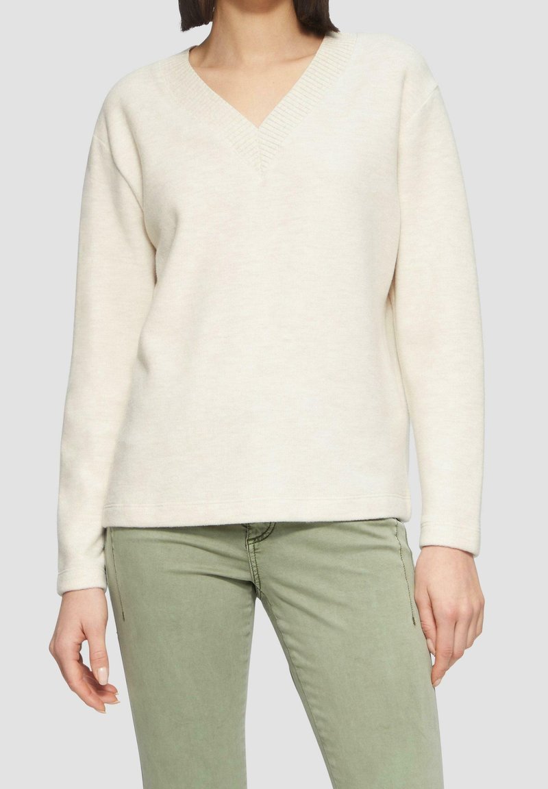 Femme portant un pull à col en V couleur crème et un pantalon vert clair devant un fond uni.