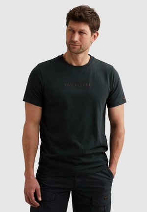 Muž nosiaci čierne tričko PME Legend s krátkym rukávom a tmavé cargo nohavice, stojaci s jednou rukou v vrecku, pozerajúci sa mierne na stranu.